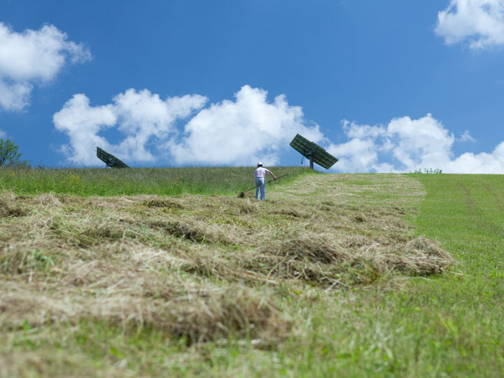 Mann beim händischen Heuen auf Wiese; Hintergrund mit Solarpanelen